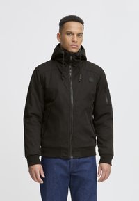 Schwarze Zip-Jacke mit Kapuze, ausgestattet mit seitlichen Taschen und einer Ärmeltasche, aus einem strapazierfähigen Material gefertigt. Gerippte Bündchen und Saum.