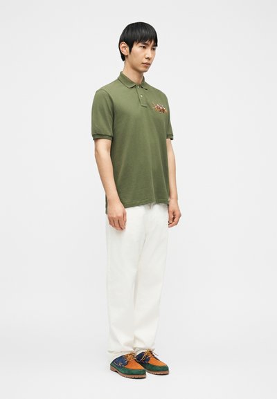 Άνδρας που φοράει πράσινο ελιά πόλο με κέντημα ιππέων, λευκό παντελόνι και πολύχρωμα δετά παπούτσια, στέκεται μπροστά σε απλό λευκό φόντο.