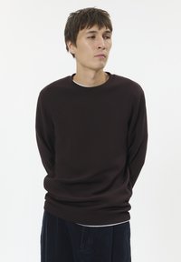Brown crewneck sweatshirt with long sleeves, made of soft material, features a relaxed fit and is layered over a white shirt.
