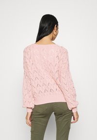 Pull en maille rose clair avec une coupe ample, présentant un motif ajouré et des poignets côtelés, accompagné d'un pantalon cargo vert olive.