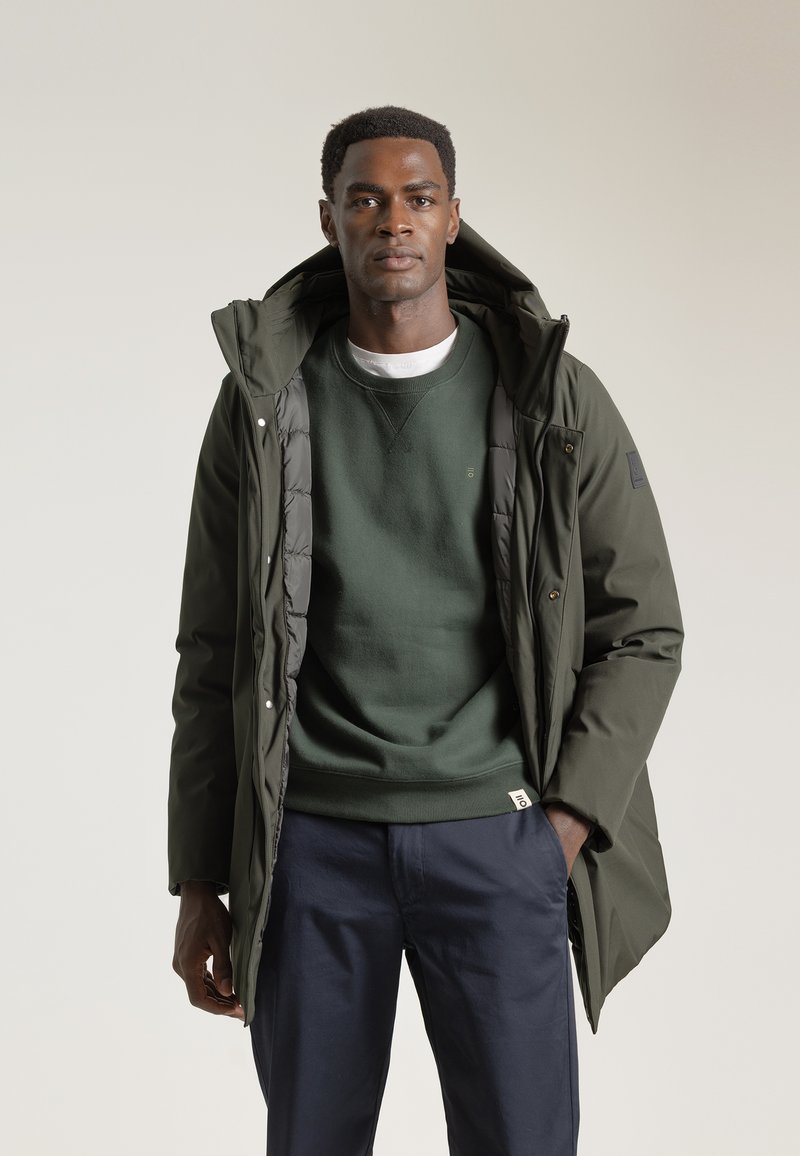 Chaqueta acolchada verde oscuro con capucha, con cierre de cremallera y botones a presión, llevada sobre un sudadera verde a juego y pantalones marinos.