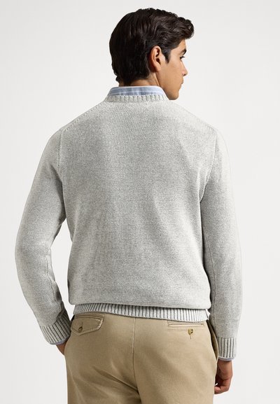 Svetlo sivi pleteni pulover z rebrastimi manšetami in spodnjim robom, z okroglim izrezom, nošen prek svetlo modre srajce z ovratnikom, skupaj z bež hlačami.