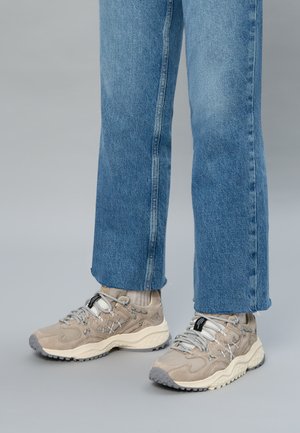 Person, der hellbeige Sneaker mit grauen Akzenten und weißen Sohlen trägt, kombiniert mit hellbeigen Socken und blauen Jeans.