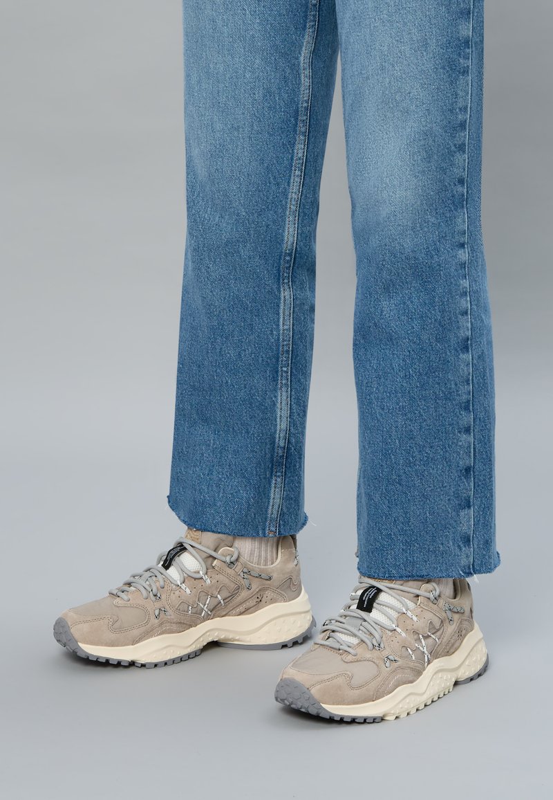 Osoba nosząca jasnobeżowe trampki z szarymi akcentami i białymi podeszwami, w zestawieniu z jasnobeżowymi skarpetami i niebieskimi dżinsami.