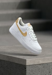 White Nike Air Force 1 sneaker with a gold swoosh, featuring a smooth leather upper, perforated toe box, and textured rubber sole.