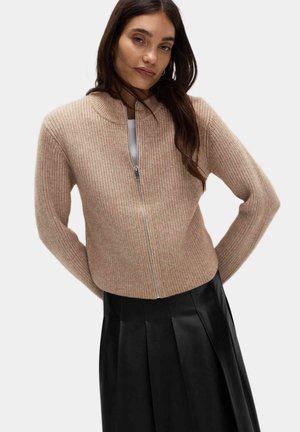 Ribbede strikcardigan i lys beige med lynlås foran og høj krave. Parret med en sort plisseret nederdel.