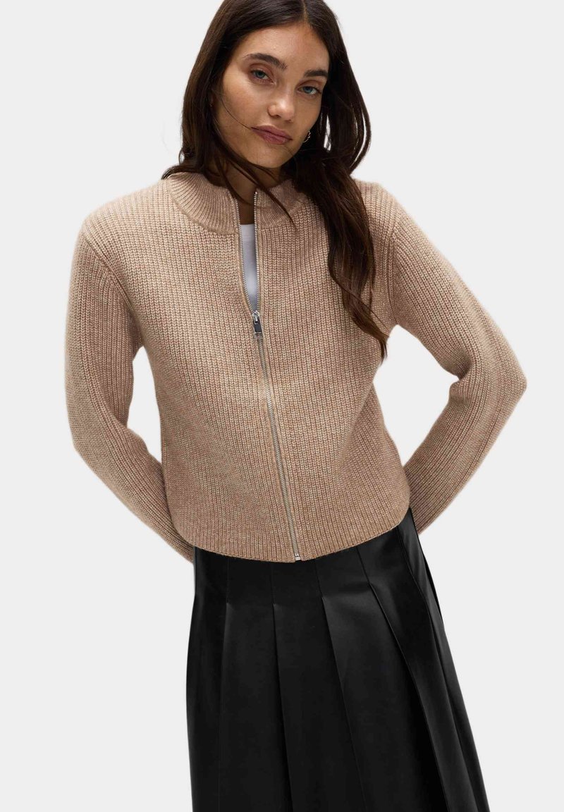 Hladce pletený kardigan ve světle béžové barvě s předním zipem a vysokým límcem. V kombinaci s černou skládací sukní.