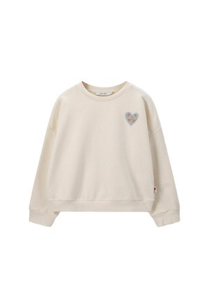 Sweatshirt couleur crème avec un col rond, des épaules tombantes et un patch en forme de cœur avec un motif floral sur la poitrine gauche. Texture douce.