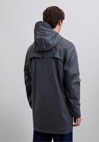 Joven con una larga chaqueta impermeable gris oscuro con capucha adjunta, visto desde atrás sobre un fondo claro y liso.