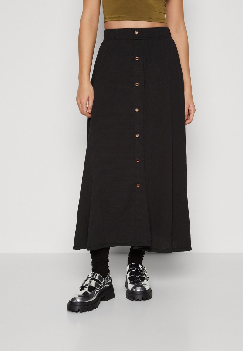 Femme portant une jupe midi noire à boutons sur le devant, des chaussettes noires, des chaussures à plateforme épaisses argentées et un crop top vert olive.