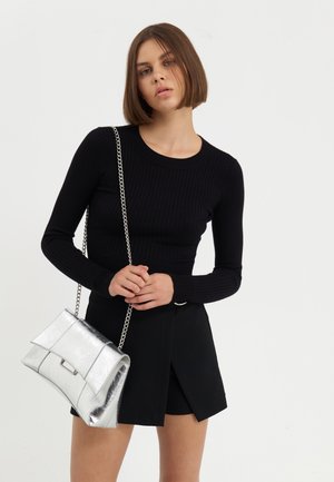 Jeune femme aux cheveux bruns courts portant un haut noir à manches longues et une jupe noire, tenant un sac à main argenté métallisé avec une bandoulière chaîne.