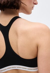 Γυναίκα με κοντά καστανά μαλλιά που φοράει μαύρο αθλητικό σουτιέν τύπου racerback με το λευκό ελαστικό λάστιχο που φέρει την επιγραφή "BJÖRN BORG".