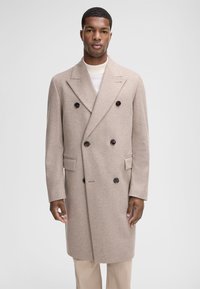 Cappotto in lana beige a doppiopetto con grandi bottoni, tasche laterali e revers a punta, caratterizzato da una vestibilità su misura e un taglio di lunghezza media.