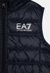 Mornarica pufasta brezrokavnik s kvadratnim vzorcem, dolgi zadrgo in logotipom "EA7 EMPORIO ARMANI" v srebrni barvi. Gladek, sijoč material.