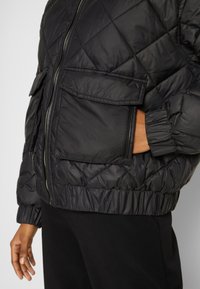 Czarna kurtka puchowa z pikowaną fakturą, z ściągaczami przy mankietach i u dołu, wyposażona w dwie przednie kieszenie oraz centralny zamek. Gładki, błyszczący materiał.