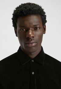 Schwarzes Samthemd mit spitzem Kragen, verziert mit glänzenden Knöpfen und einer glatten Textur, getragen von einer Person mit dunklem, lockigem Haar.