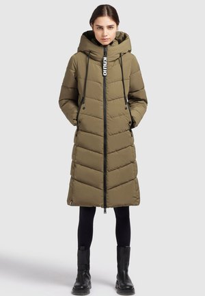 Frau trägt knie langen olivgrünen, gesteppte Steppmantel mit Kapuze, schwarze Leggings und schwarze Stiefeletten, steht vor einfachem Hintergrund.