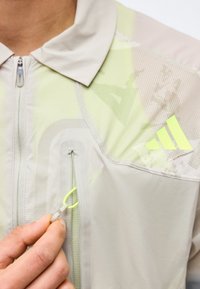 Main tenant une tirette de fermeture éclair sur une veste beige clair avec un logo Adidas jaune néon sur la poitrine près d'une poche zippée.