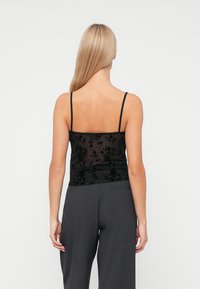 Top in pizzo floreale nero con spalline sottili e un tessuto trasparente, abbinato a pantaloni grigi con dettaglio sulla tasca posteriore.