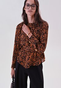 Blusa stampa leopardo arancione e nera con maniche lunghe, scollatura arricciata e dettaglio peplum laterale, abbinata a pantaloni neri.