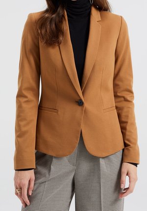 Femme portant un blazer camel sur un col roulé noir et un pantalon à carreaux gris, avec des bagues à la main gauche.