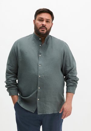 Mężczyzna z brodą, ubrany w zieloną koszulę z kołnierzykiem mandarynkowym zapinaną na guziki oraz ciemne spodnie na jednolitym białym tle.