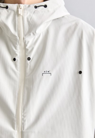 Chaqueta cortavientos blanca con cierre de cremallera, capucha con cordón, dos botones a presión negros y un pequeño logo A C W en la parte frontal.