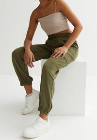 Oliwkowe spodnie dresowe z mankietami, elastycznym paskiem i bocznymi kieszeniami, w połączeniu z białymi trampkami i jasnobrązowym krótkim topem.