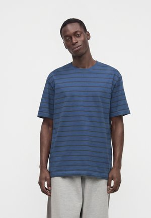 Homme portant un t-shirt bleu à manches courtes avec des rayures horizontales foncées et un pantalon gris clair, debout devant un fond blanc uni.