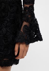 Melnas kleitas ar acs audumu, izceļot izceltas ziedu aplikācijas, plati piedurkņu un teksturētu virsmu. Tuvplāns, kurā redzama roka, kas tur kleitu.