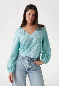 Lyseblå bluse med tekstureret blomsterbroderi, V-hals og snøredetaljer. Puffede ærmer med elastiske manchetter kombineret med denim jeans.