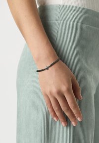 Bracelet noir tressé avec un accent en croix métallique, porté sur une main contre un fond de tissu en velours côtelé vert clair texturé.