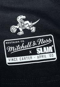 Un tessuto nero presenta una grafica bianca di un dinosauro in abbigliamento da basket che tiene una palla, con il logo "Mitchell & Ness x SLAM®" sotto.