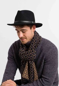 Homme portant un chapeau fedora noir, une écharpe à motifs sombres et un pull en laine gris texturé, regardant vers le bas avec les mains jointes.