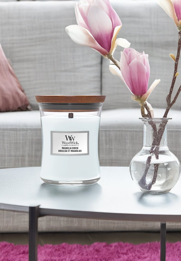 Candela WoodWick in un barattolo di vetro trasparente con coperchio in legno, cera bianca e un'etichetta. Accompagnata da un vaso di vetro con fiori di magnolia.