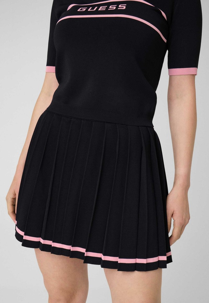 Gros plan d'une personne portant une jupe plissée noire et un haut noir assorti avec un liseré rose et le logo "GUESS" sur la poitrine.