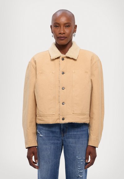 Veste en denim beige raccourcie avec un col doux, deux poches avant et des boutons en argent ; portée sur un jean bleu déchiré.