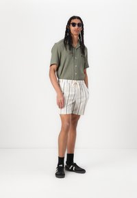 Chemise olive à manches courtes, shorts légers rayés avec taille élastique et baskets noires avec accents blancs. Lunettes de soleil noires portées.