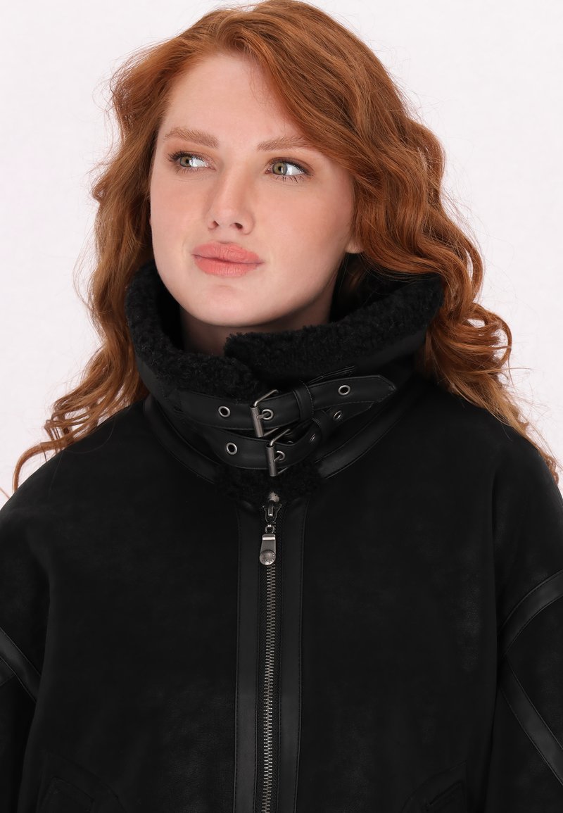 Schwarze Wildlederjacke mit hohem Kragen und abnehmbarer, plüschiger Innenfütterung, verstellbaren Riemen und einem Frontreißverschluss. Glatte Textur mit minimalen Akzenten.