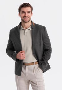 Chaqueta gris oscuro con cuello alto y detalles en cuero marrón, llevada sobre un polo beige claro. Cinturón beige con hebilla rectangular y pantalones claros.