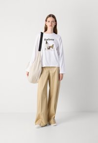 Chemise blanche à manches longues avec un graphisme "Barbour", pantalon beige à jambes larges et un sac bandoulière crème. Des baskets blanches complètent la tenue.