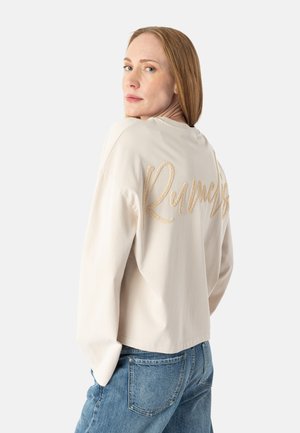 Kvinde med glat rødt hår iført en beige langærmet top med "Runmes" broderet på ryggen, kombineret med blå jeans.
