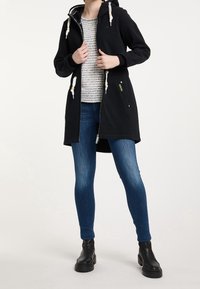 Personne portant une veste à capuche noire zippée avec des cordons blancs, un haut blanc texturé, un jean skinny bleu, et des bottines noires.