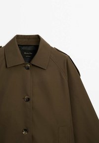 Ruskea trench-takki, jossa on klassinen kaulus, edessä napitus ja olkapäällysteet. Valmistettu sileästä kankaasta, joka on muotoiltu istuvaksi.