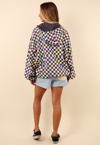 Veste à carreaux colorée en rose, bleu, jaune et noir avec une capuche, portée sur un short en jean clair et des baskets noires. Tissu texturé.