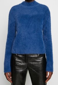 Pull bleu en maille douce avec col montant et manches longues, associé à un pantalon noir ajusté en simili cuir brillant à texture crocodile.