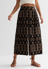 Wide-leg black pants with brown geometric patterns worn with brown strappy sandals and a white crochet crop top.