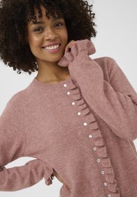 Mosolygó nő göndör hajjal, rózsaszín kötött pulóvert visel gyöngygombokkal és fodros mandzsettával, keze az állához ér, világos háttér.