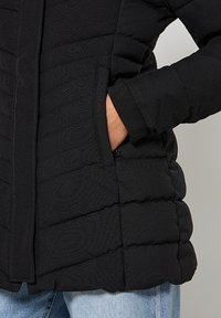 Veste noire matelassée avec texture quiltée verticale, dotée d'une fermeture éclair frontale et de poches latérales, fabriquée en matériau lisse et chaud.