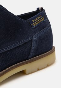 Pantof de velur bleumarin cu un finisaj texturat, cusături în contrast, o talpă din cauciuc bej și un eticheta albastră inscripționată "bugatti."